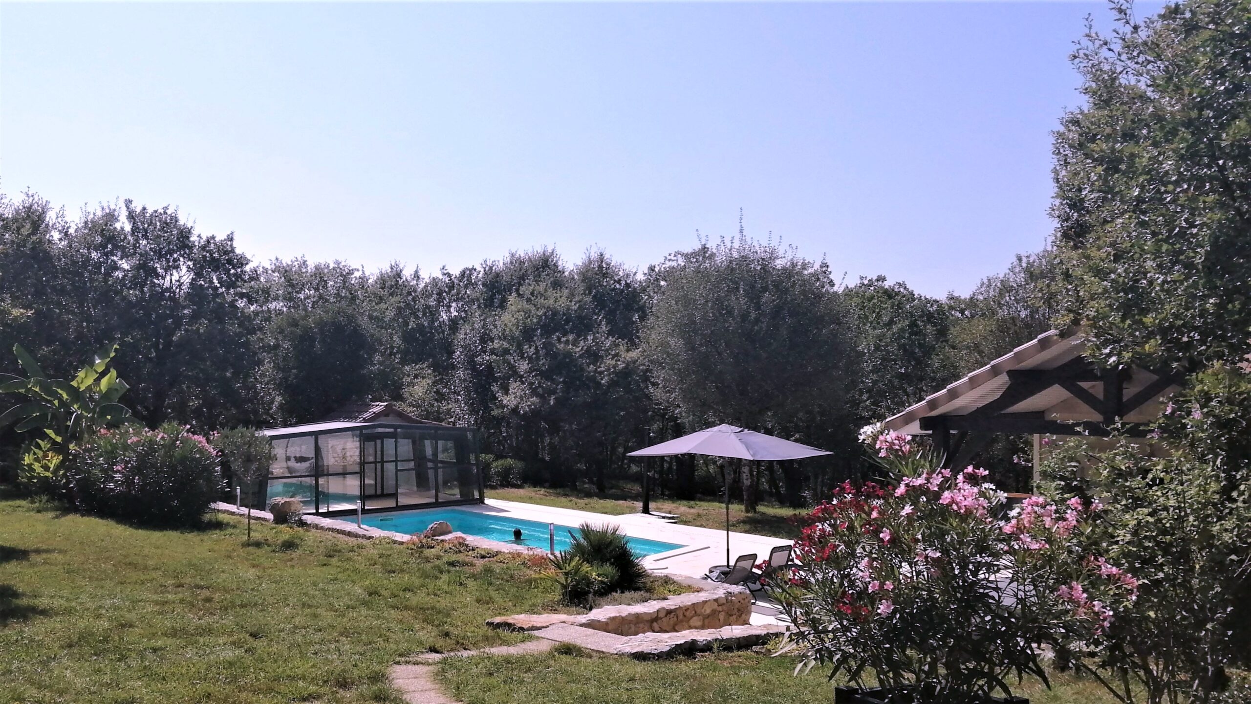 Vue d’ensemble de la piscine du gîte naturiste DACANIRO avec l’abri replié, son environnement et une personne qui se baigne