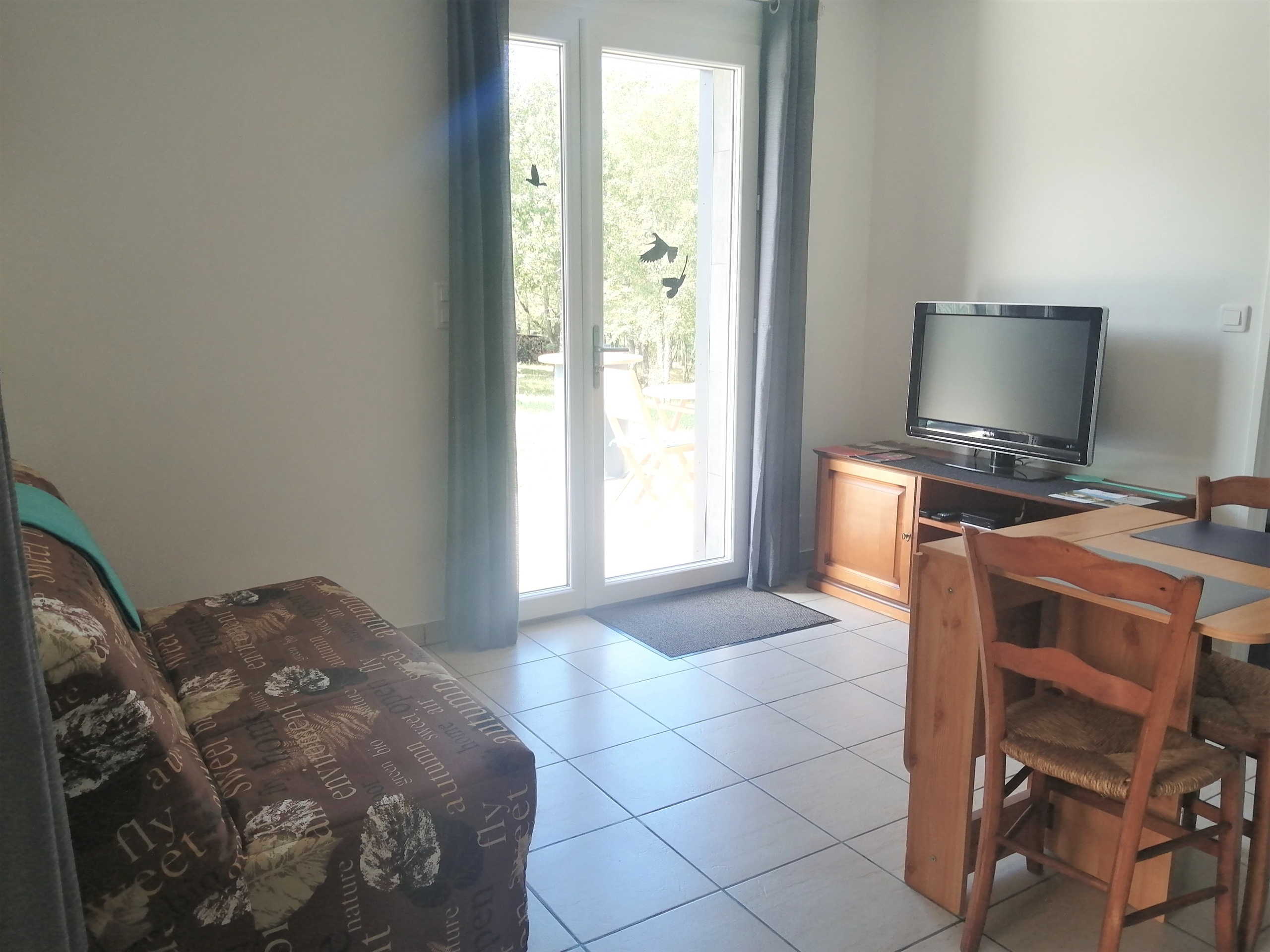 Pièce à vivre du gîte naturiste DACANIRO avec canapé BZ, meuble TV, table, chaises et vue sur le jardin