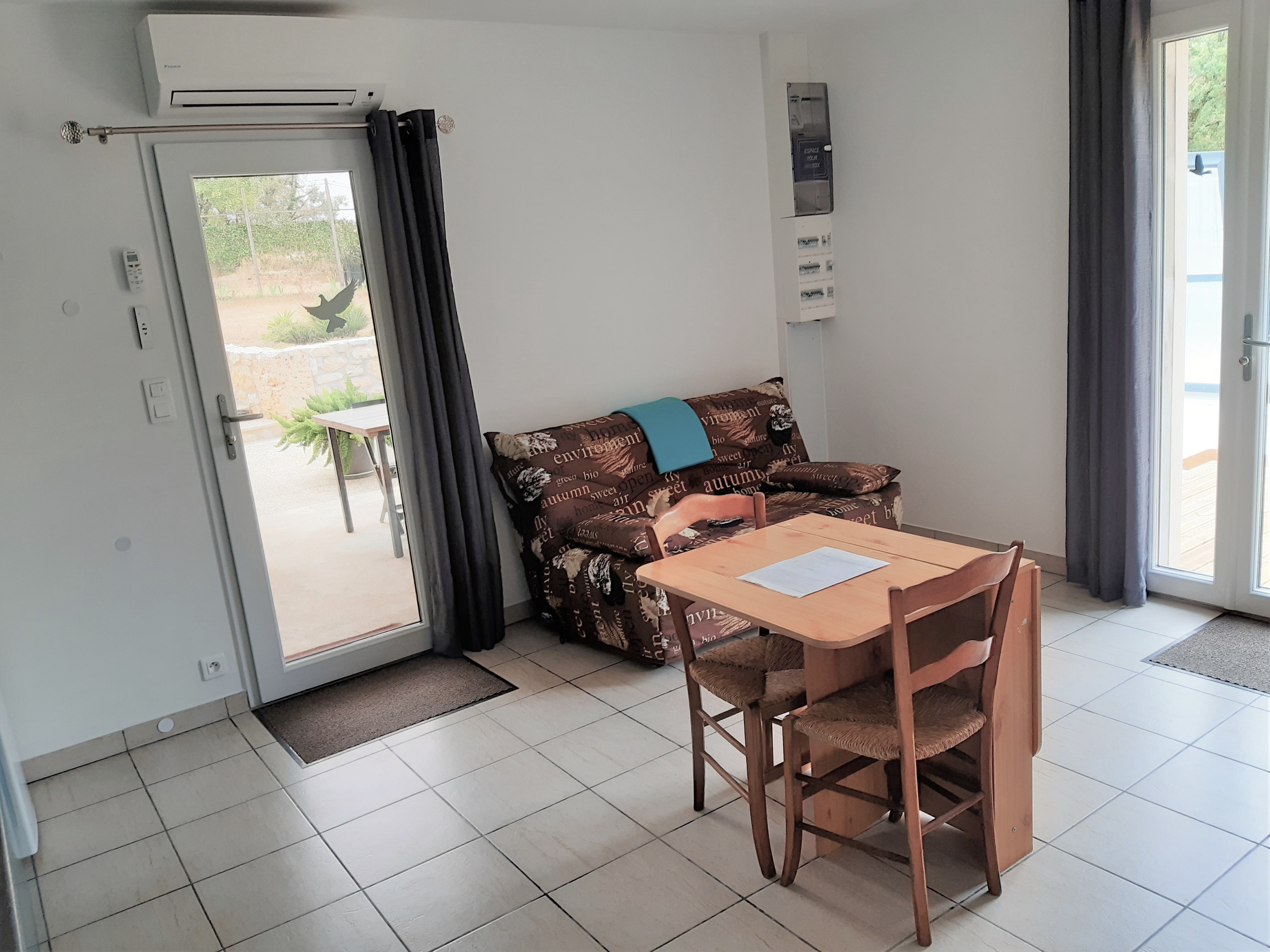 Pièce à vivre du gîte naturiste DACANIRO avec le canapé BZ, la table et la porte‑fenêtre donnant sur la terrasse couverte